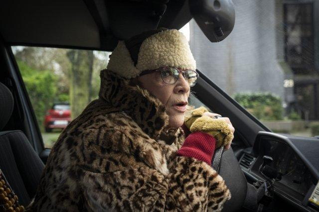 Eine ältere Frau in einem Auto, mit Pelzmütze und -mantel. Sie fährt, mit Brille, nach vorne schauend. Ein rotes Auto ist draußen sichtbar.