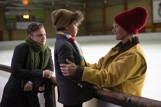 Drei Personen auf einer Eisbahn. Ein Kind und eine Frau umarmen sich. Ein Mann beobachtet von der Seite. Alle tragen Winterkleidung, Mützen und Schals.