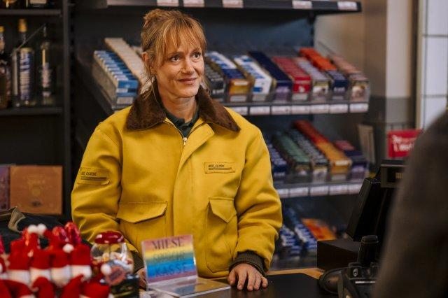 Eine Frau in einer gelben Jacke steht hinter einem Tresen mit Zigaretten. Regale mit Produkten im Hintergrund. Ein Schild mit der Aufschrift 'Kasse' befindet sich davor.