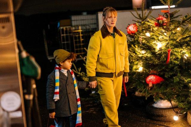Eine Frau in Gelb und ein Kind in einem Mantel halten Hände. Im Hintergrund steht ein geschmückter Weihnachtsbaum.