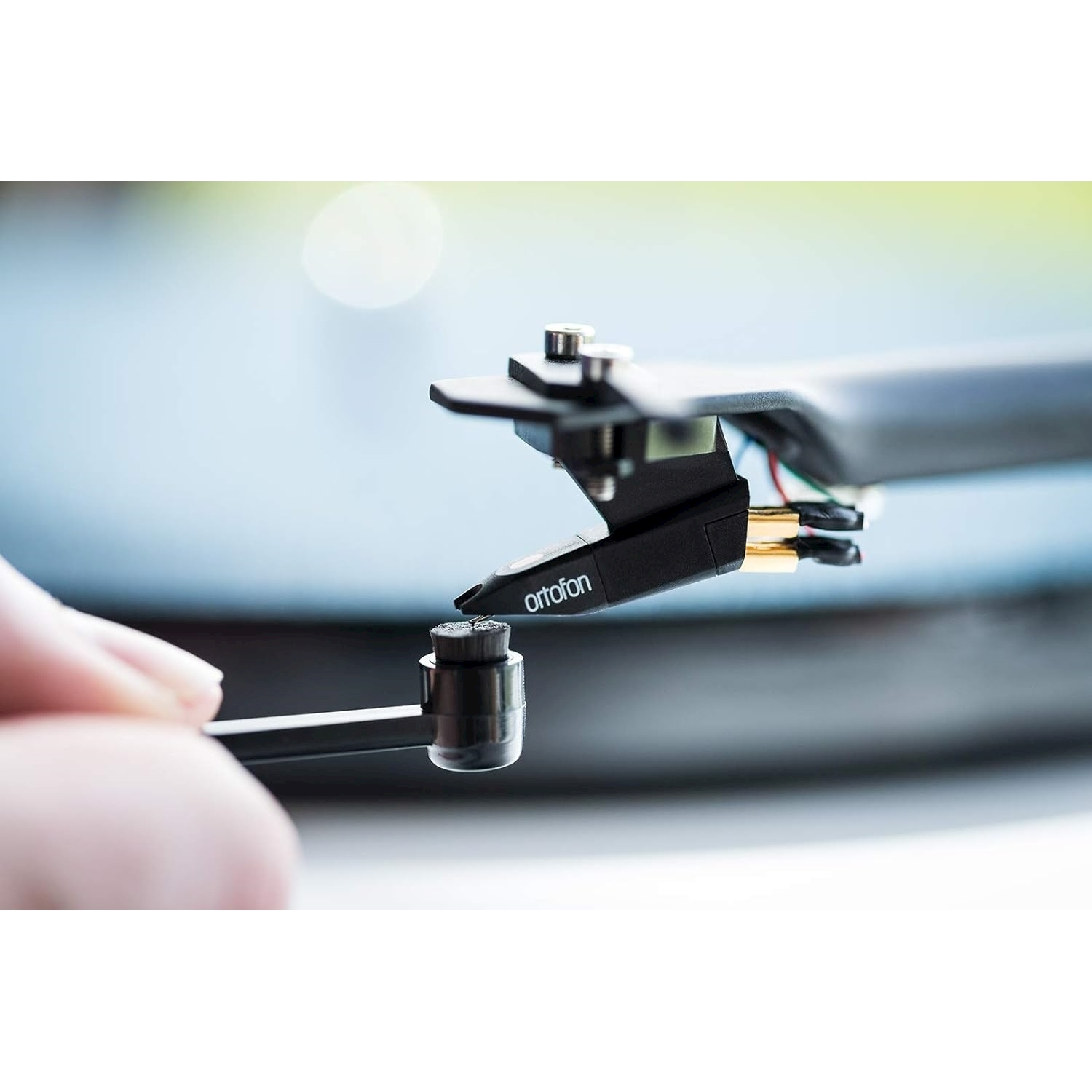 Primo piano dello stilo di un giradischi con il marchio 'ortofon' visibile. Una mano tiene un oggetto nero vicino allo stilo.