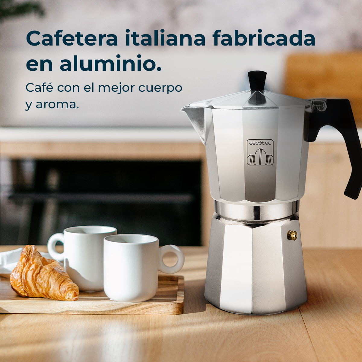 Una cafetera italiana de aluminio con asa negra, dos tazas blancas y un cruasán en una bandeja de madera.