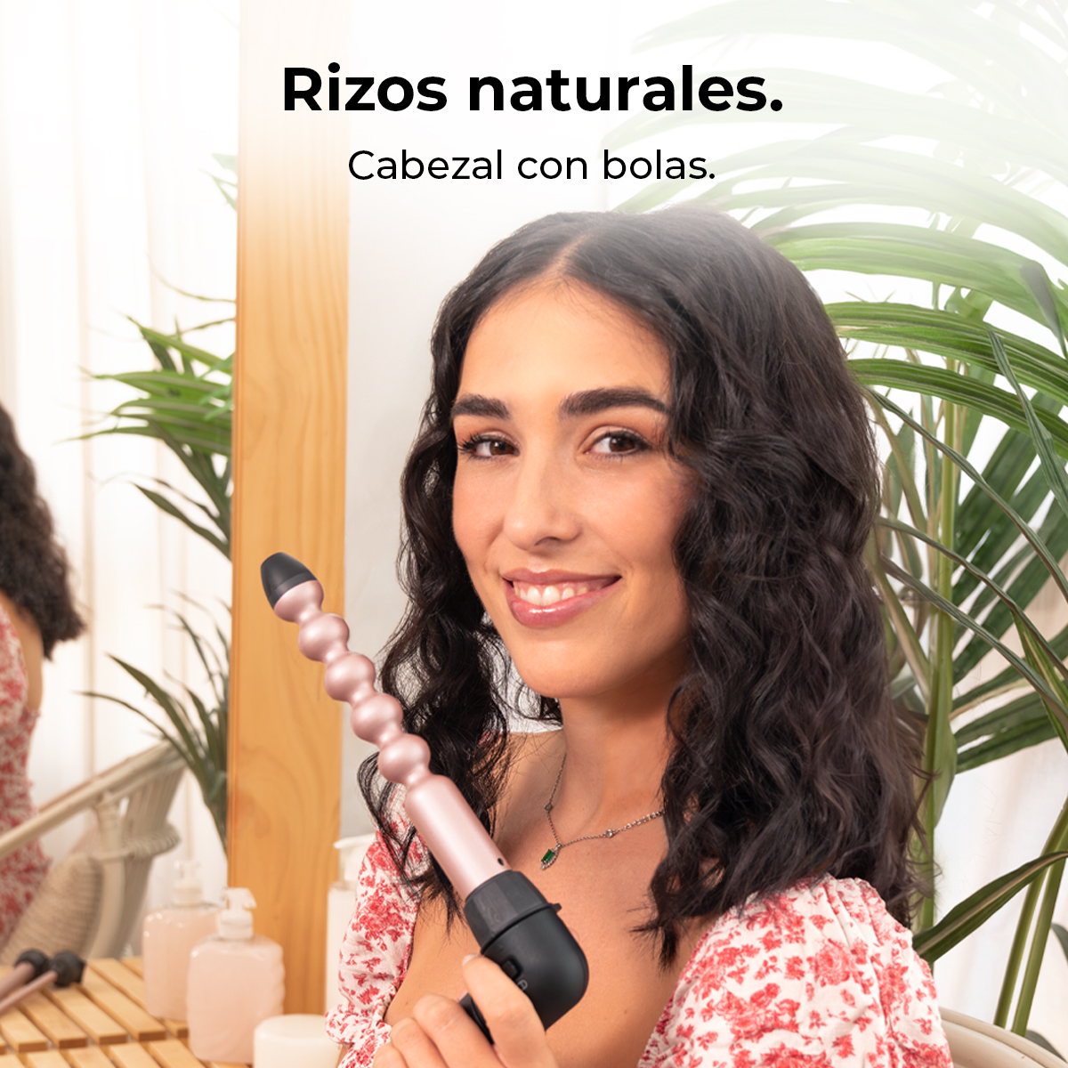 Mujer sonriente sosteniendo un rizador, con cabello rizado natural. En el fondo, un espejo, una planta y algunos productos de belleza.