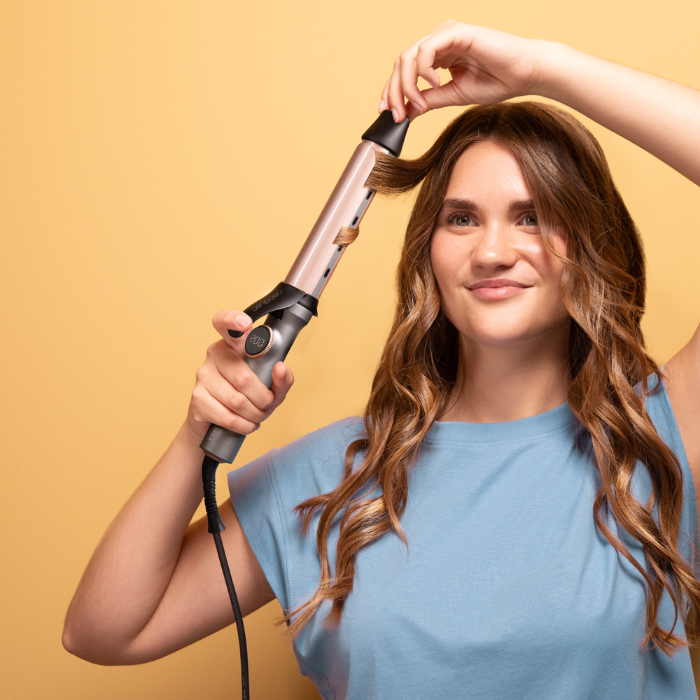 Una mujer se riza el pelo con un rizador, sonriendo. Lleva una camisa azul sobre un fondo amarillo.