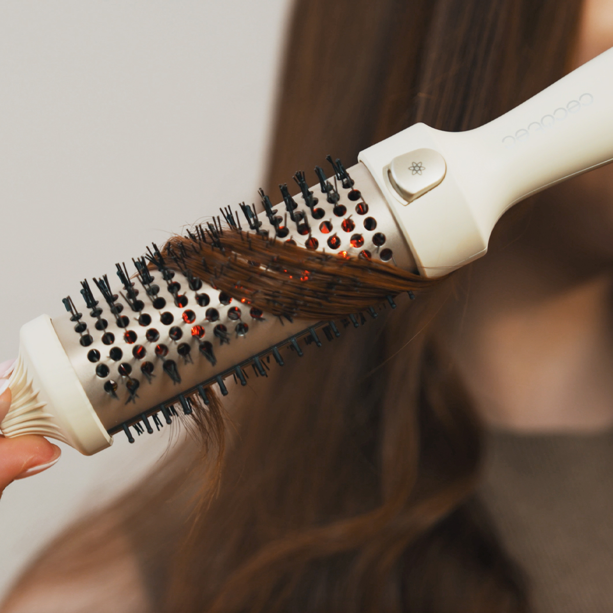 Una persona usa un cepillo de peinado beige y negro en su cabello castaño. El cepillo está cerca del cabello con un fondo neutro.