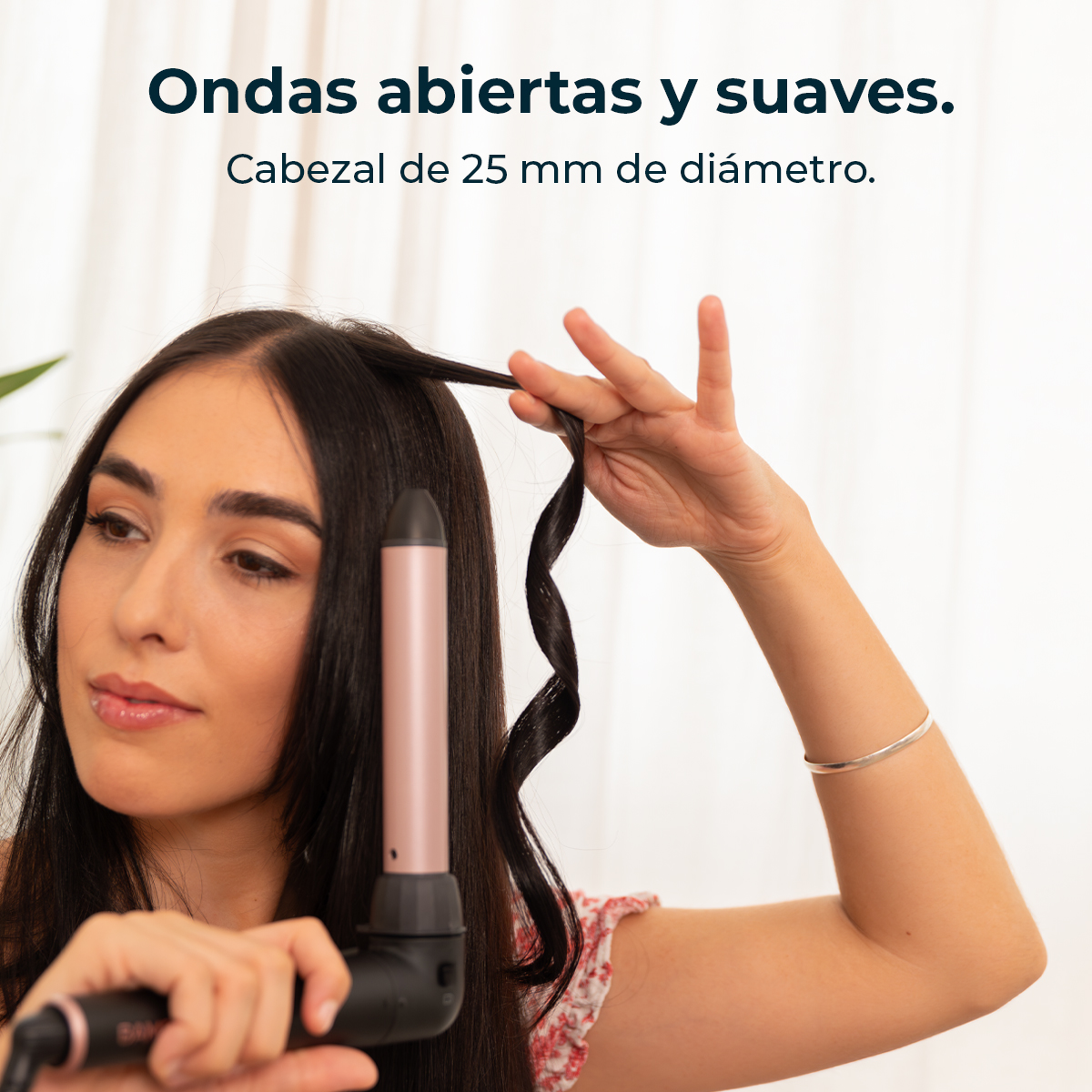 Mujer rizando su cabello oscuro con un rizador. Lleva una blusa floral y una pulsera plateada.