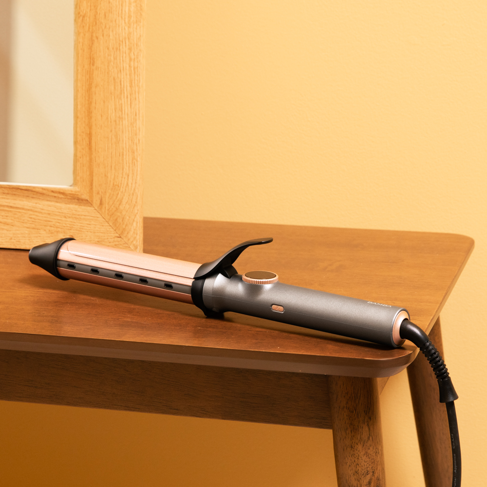 Una plancha para el pelo descansa sobre una mesa de madera. El fondo es una pared amarilla. Un marco de espejo está a la izquierda.