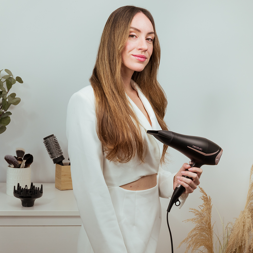 Mujer de blanco sosteniendo un secador de pelo negro. Herramientas para el cabello sobre una mesa blanca. Fondo blanco.