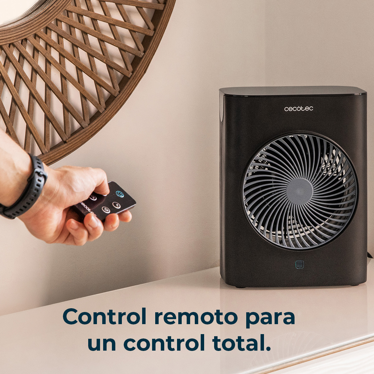 Una mano sostiene un mando a distancia para un ventilador negro. El ventilador está sobre una superficie blanca y un espejo de madera al fondo.