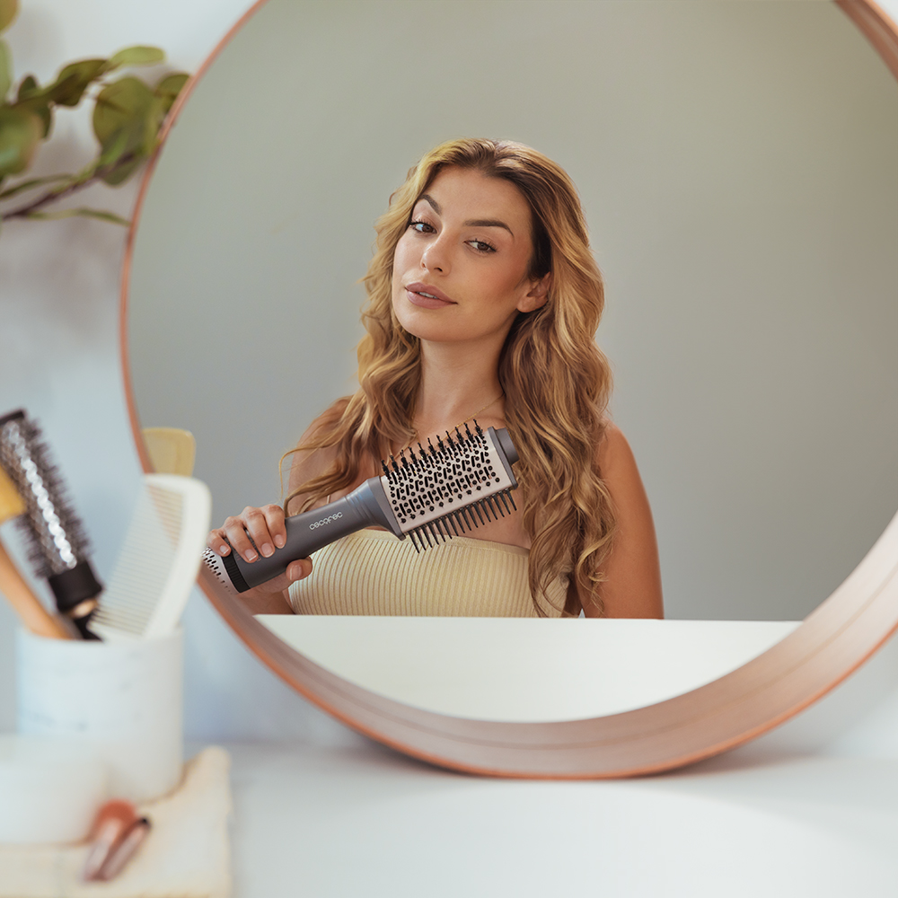 Mujer con cabello ondulado sosteniendo un cepillo para peinar el cabello frente a un espejo. Otros cepillos y vegetación son visibles.