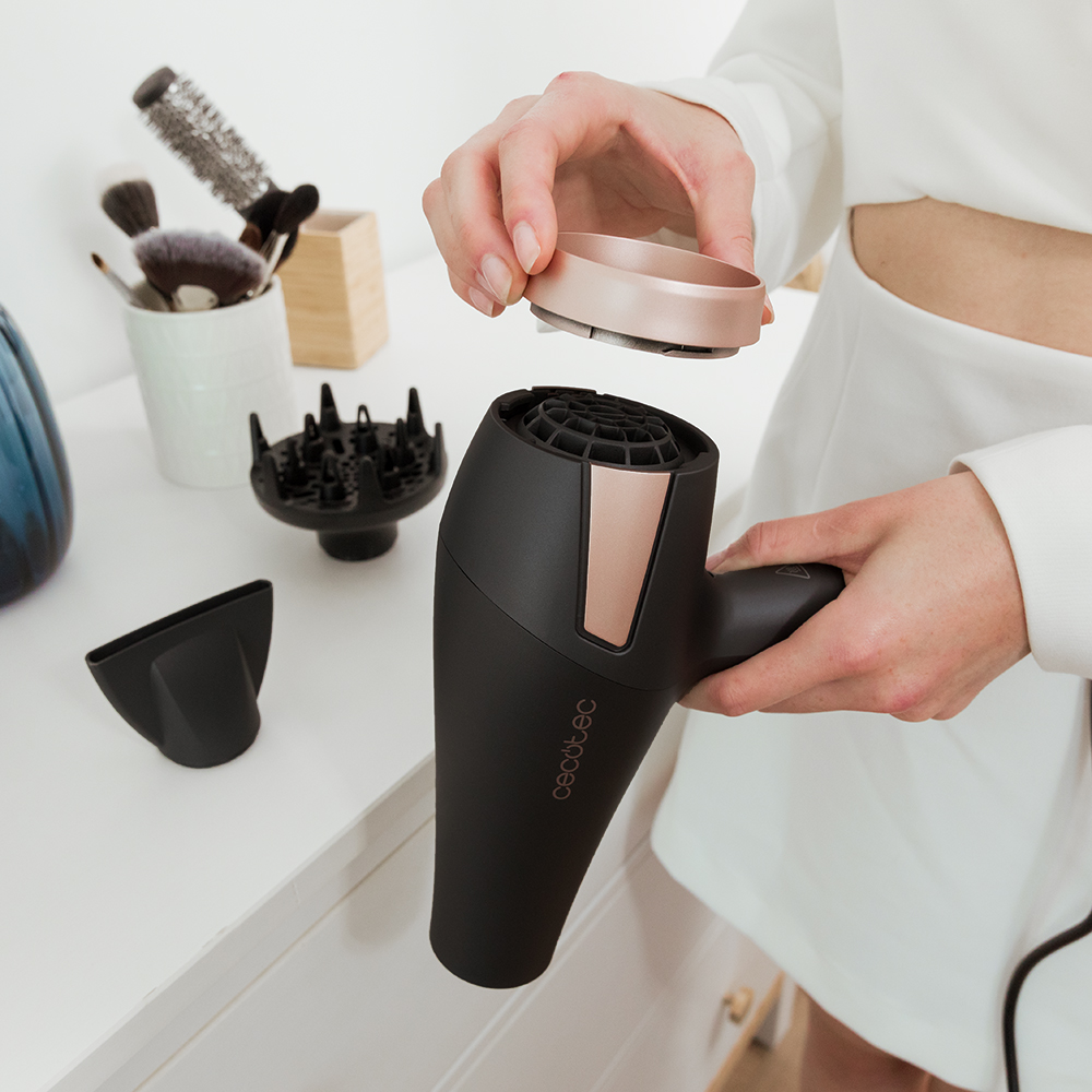 Una persona está montando un secador de pelo negro y oro rosa. Varios accesorios y productos de belleza están sobre la superficie blanca.