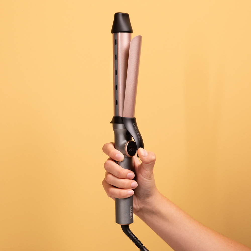 Una mano sostiene un rizador de pelo. El rizador es gris y oro rosa con pantalla digital que muestra temperatura. El fondo es amarillo.