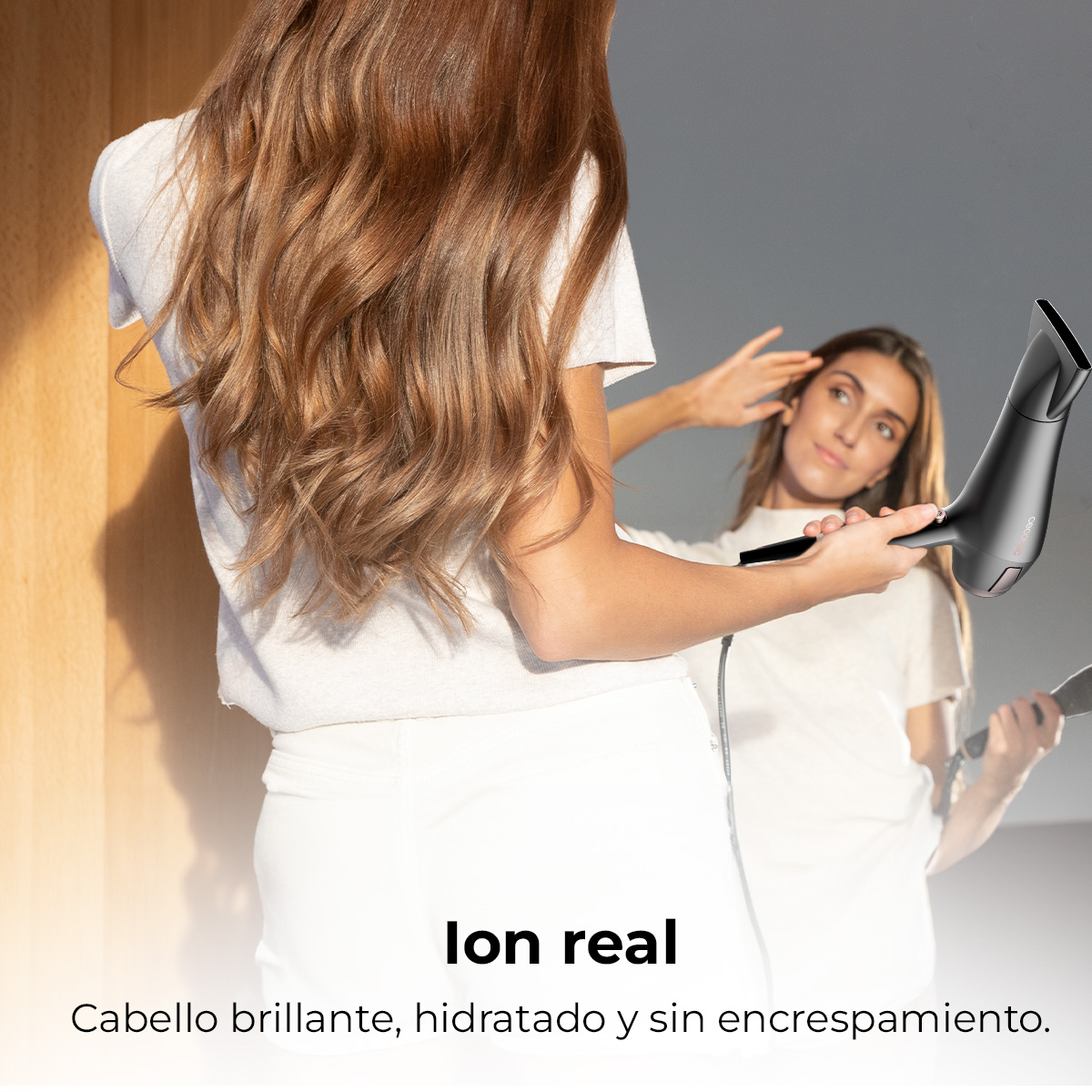 Una mujer se seca el pelo con un secador negro frente a un espejo. Otra mujer se refleja en el espejo.