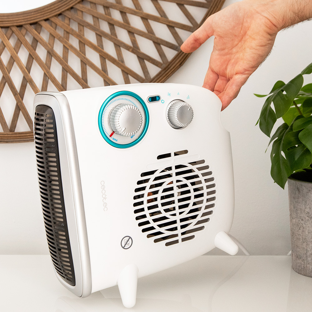 Un calentador eléctrico blanco con detalles azules y diales plateados es sostenido por una mano. Detrás hay una pieza decorativa de madera.