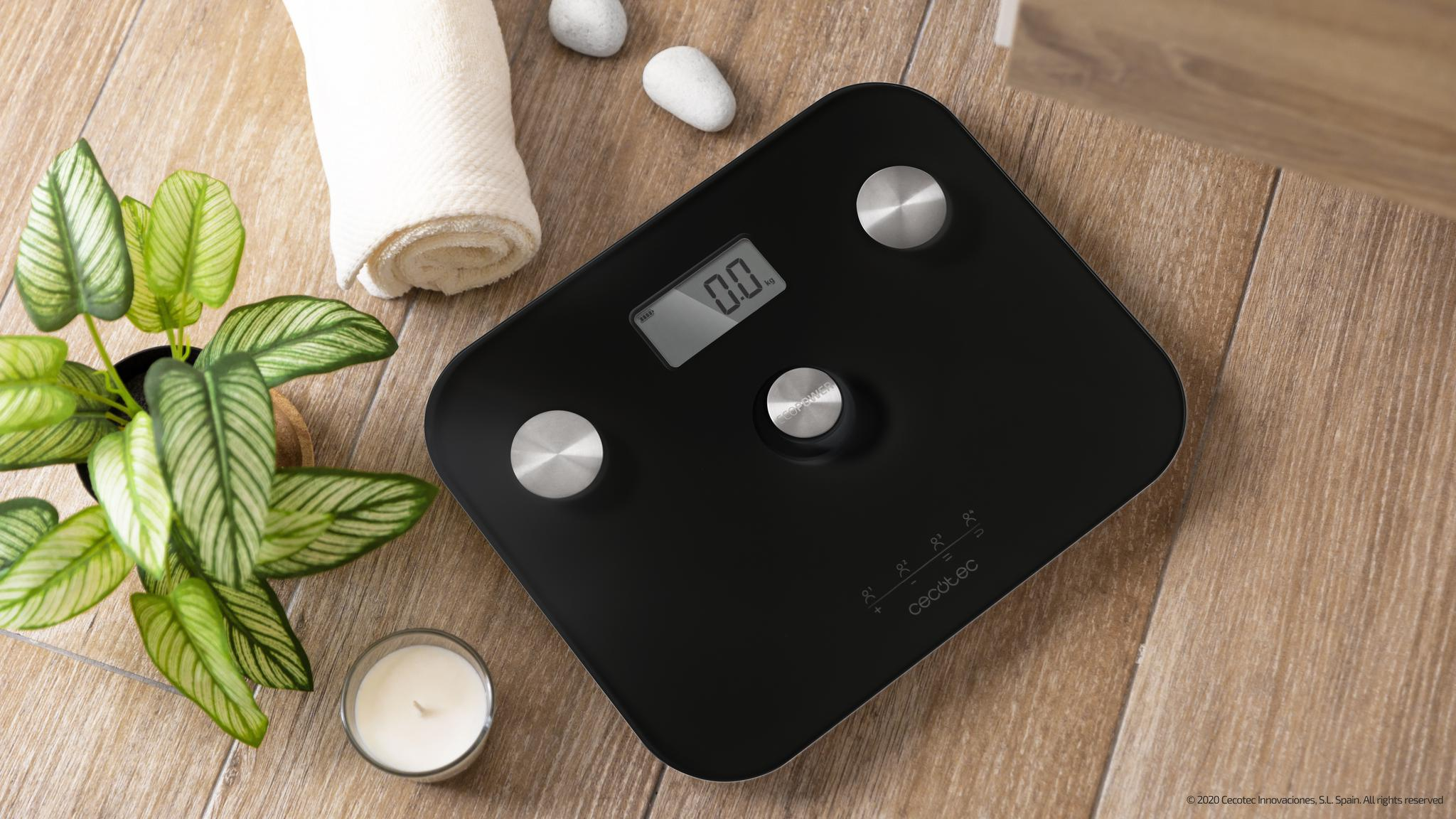 Una báscula digital negra muestra 0,0 kg, con una toalla blanca enrollada, una planta en maceta, una vela y piedras sobre una superficie de madera.