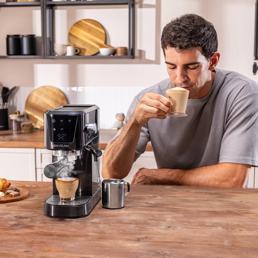 Un hombre bebe café de un vaso, junto a una cafetera. Una taza de café está sobre la máquina. Una jarra de metal está en la mesa.