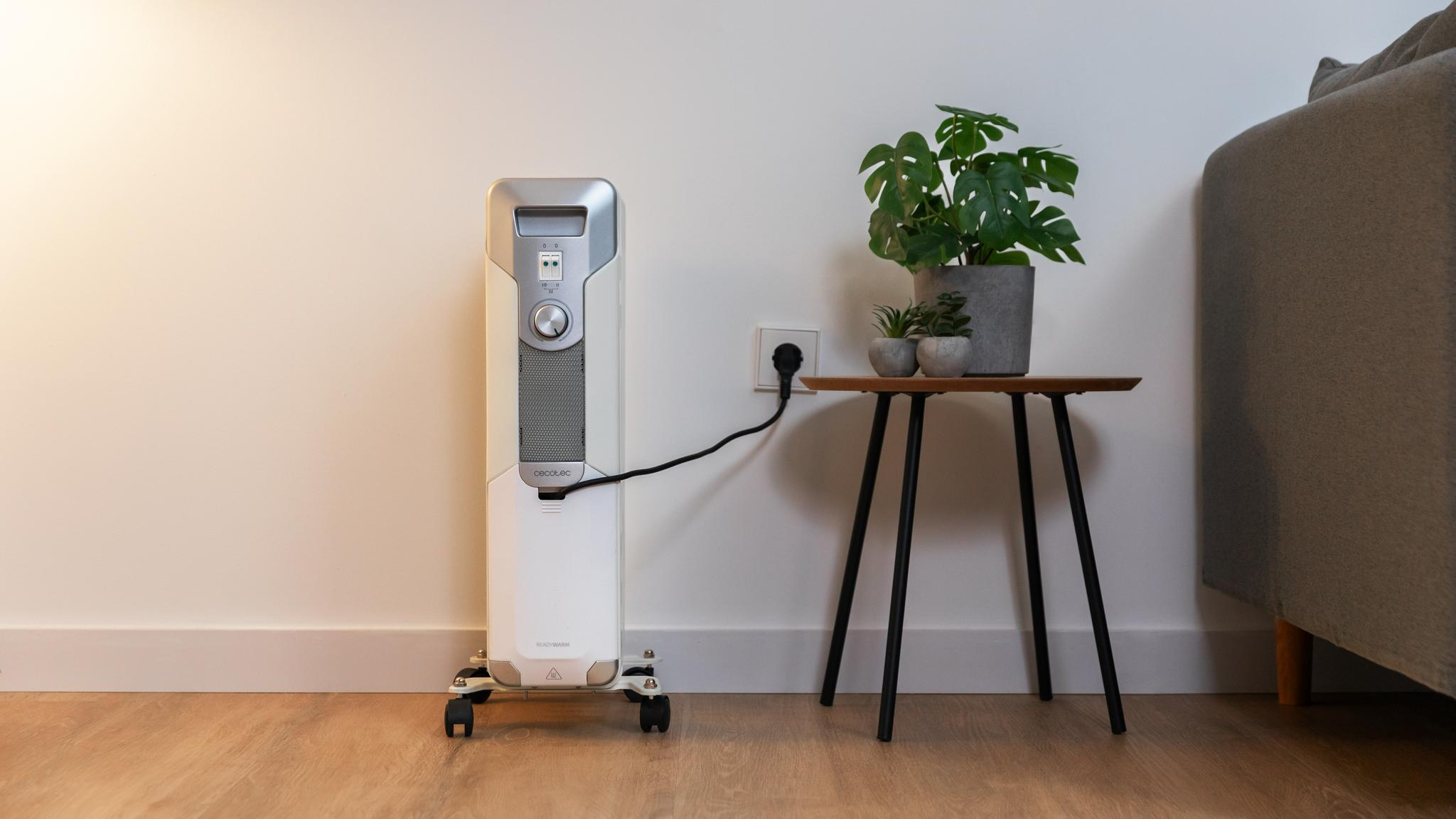 Un calentador eléctrico blanco sobre ruedas está enchufado a una toma de corriente. Una mesa pequeña sostiene plantas y un sofá a la derecha.