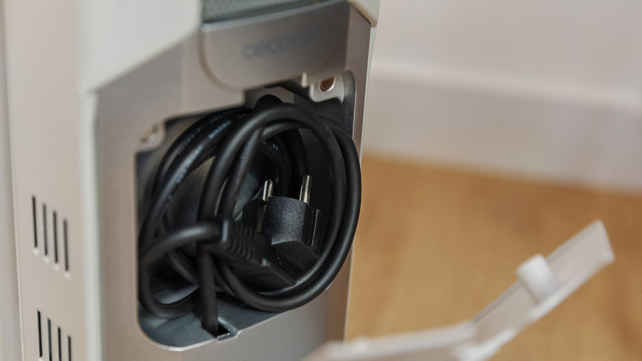 Un cable de alimentación negro con un enchufe está enrollado dentro de un compartimento de un electrodoméstico blanco y gris.