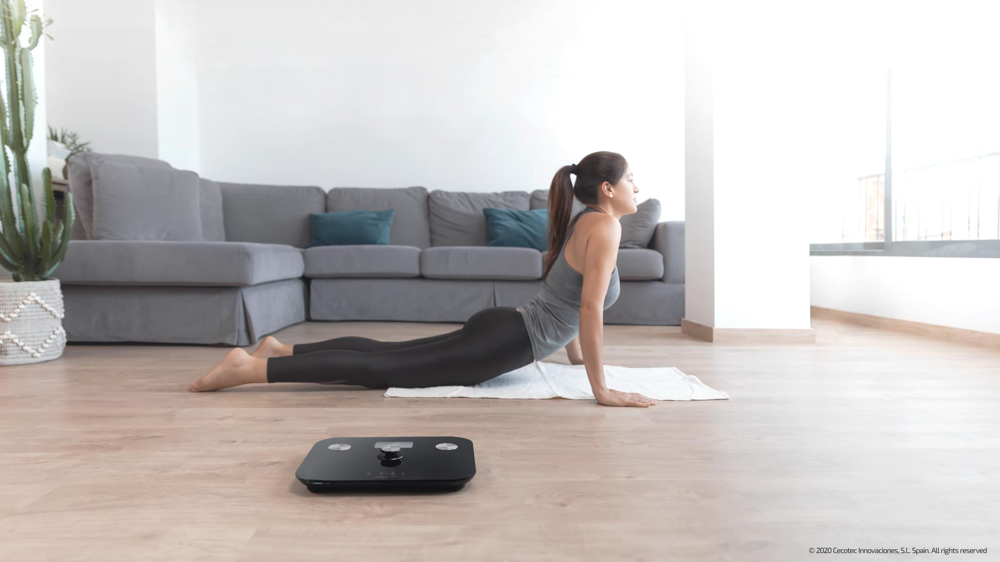 Mujer en pose de yoga en interiores, cerca de una báscula. Sofá gris, planta y ventana visibles. Mallas negras, camiseta gris, toalla blanca.