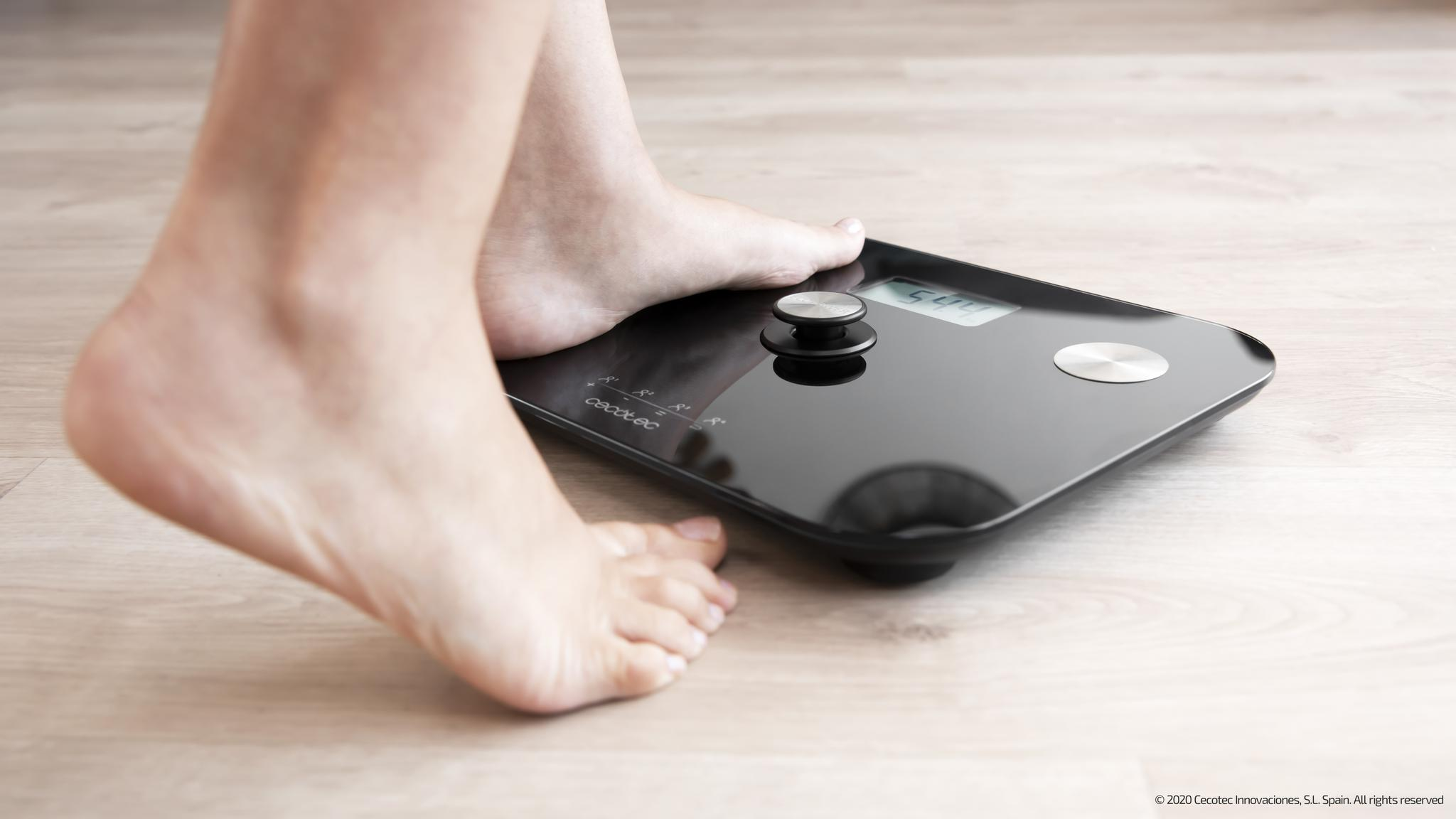 Los pies de una persona sobre una báscula digital negra, que muestra una lectura de peso. El suelo es de madera clara. La báscula está en el suelo.
