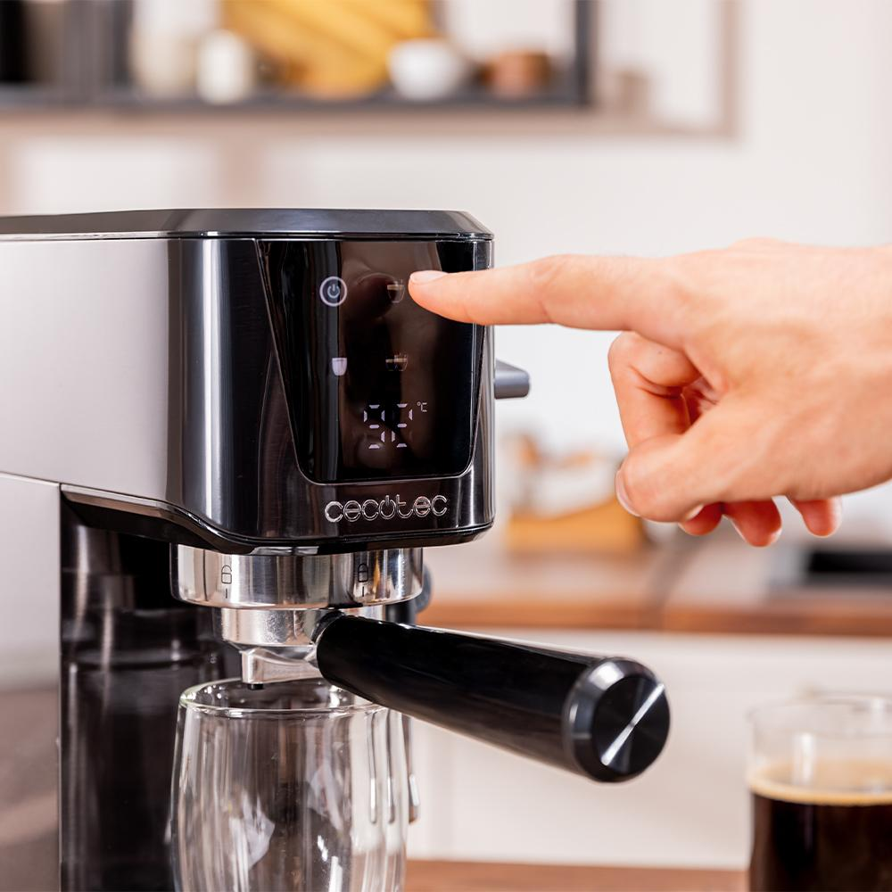 Una mano presiona un botón en una máquina de espresso negra y plateada. Hay un vaso debajo de la boquilla. Una taza de café a la derecha.
