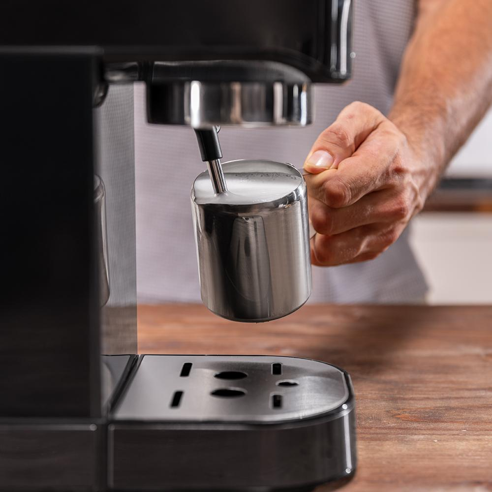 Una persona bate leche con una máquina de espresso. Una jarra de acero inoxidable se sostiene debajo de la varilla espumadora. Mesa de madera en el fondo.