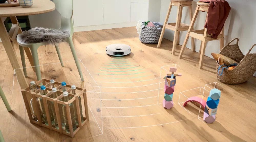 Robot sprzątający porusza się po pomieszczeniu z przeszkodami, takimi jak drewniane klocki i skrzynka z butelkami wody. Drewniana podłoga.