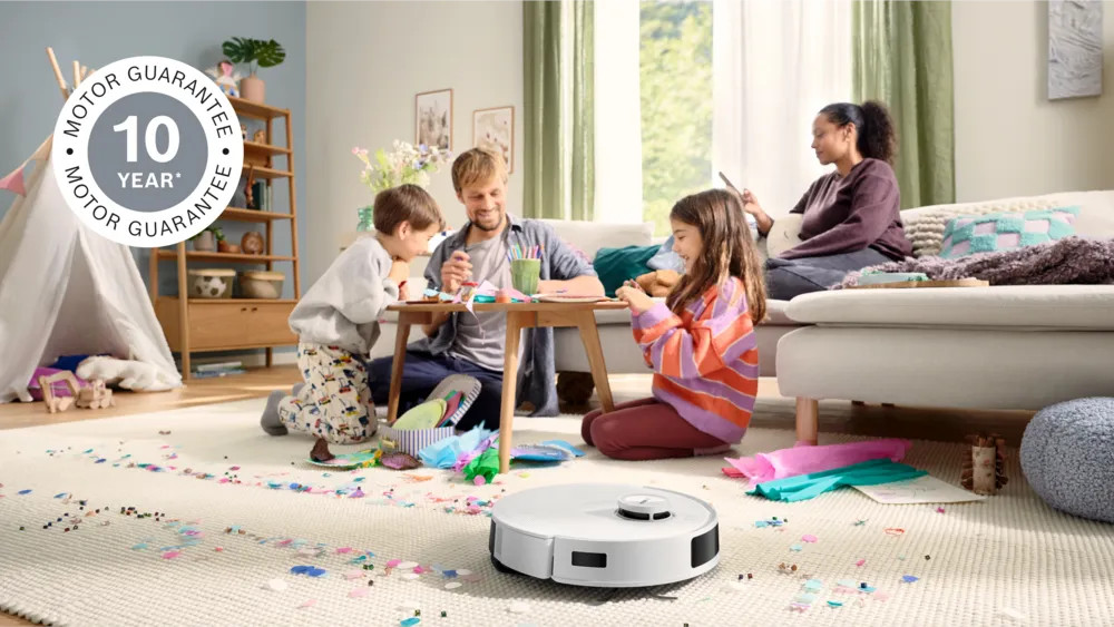 Rodzina tworzy przy stole. Biały odkurzacz robot stoi na dywanie z konfetti. Kobieta siedzi na kanapie, trzymając telefon.