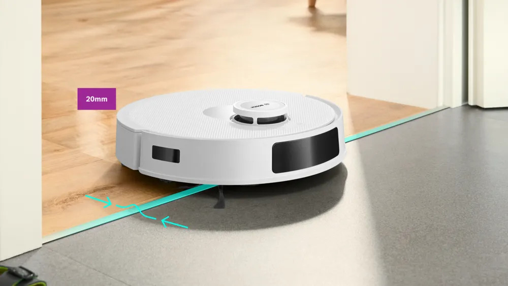 Biały robot odkurzający na drewnianej podłodze. Zbliża się do drzwi. Podłoga zmienia się w szary beton.