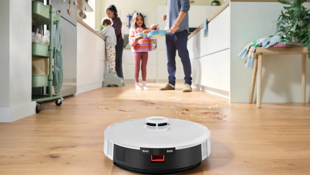 Rodzina w kuchni rozlała jedzenie. Robot odkurzacz jest na pierwszym planie. Członkowie rodziny stoją przy blacie i sprzątają.