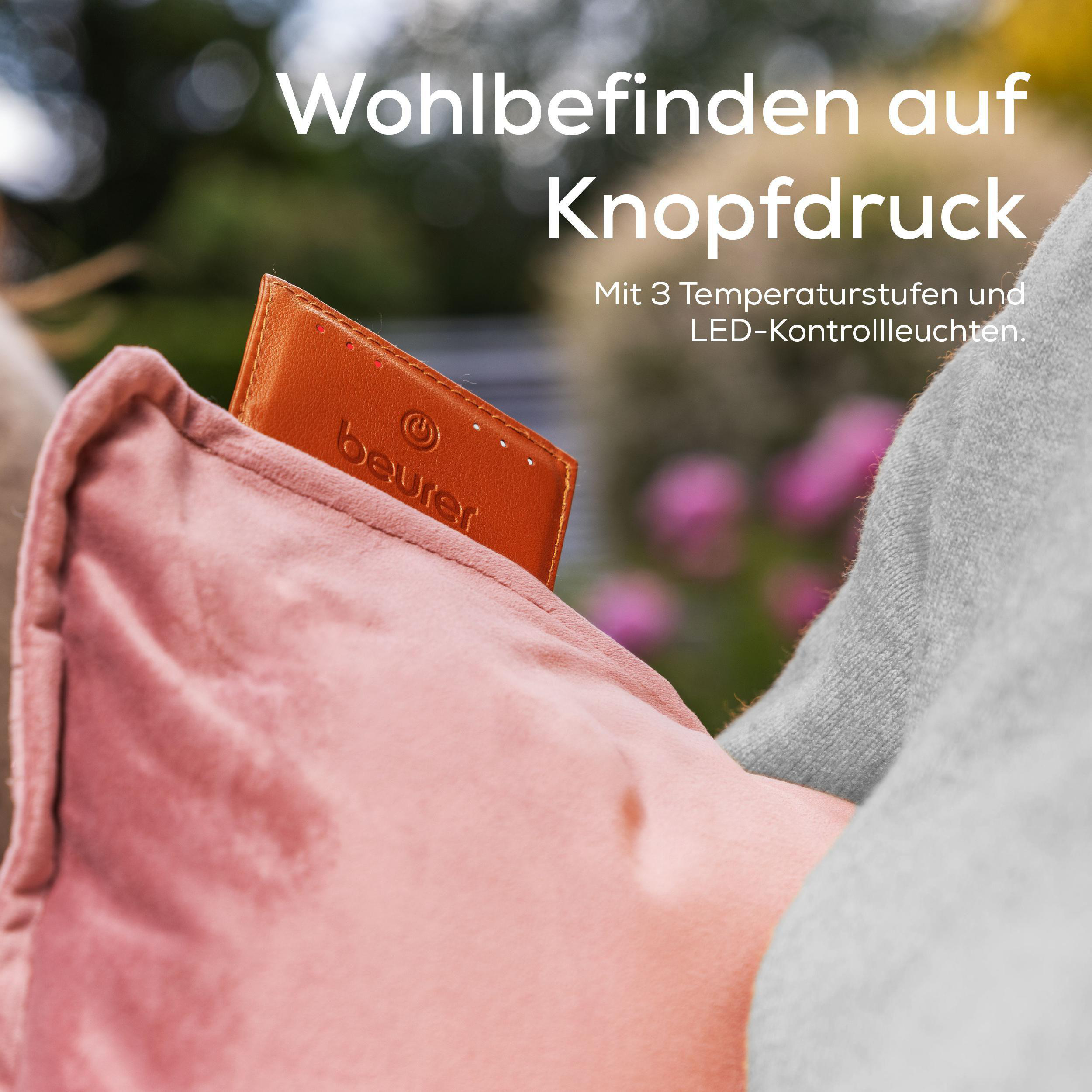 Eine Hand hält ein rosa Kissen mit einem braunen Etikett. Text auf dem Etikett und oben lautet 'Wohlbefinden auf Knopfdruck'.