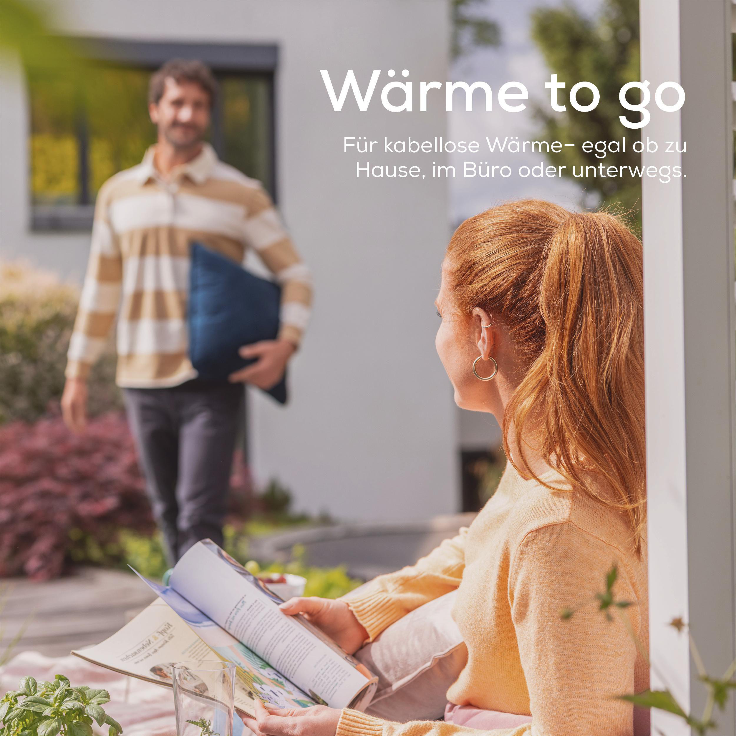 Eine Frau liest draußen ein Magazin; ein Mann kommt mit einem blauen Kissen. Text sagt 'Wärme to go'.