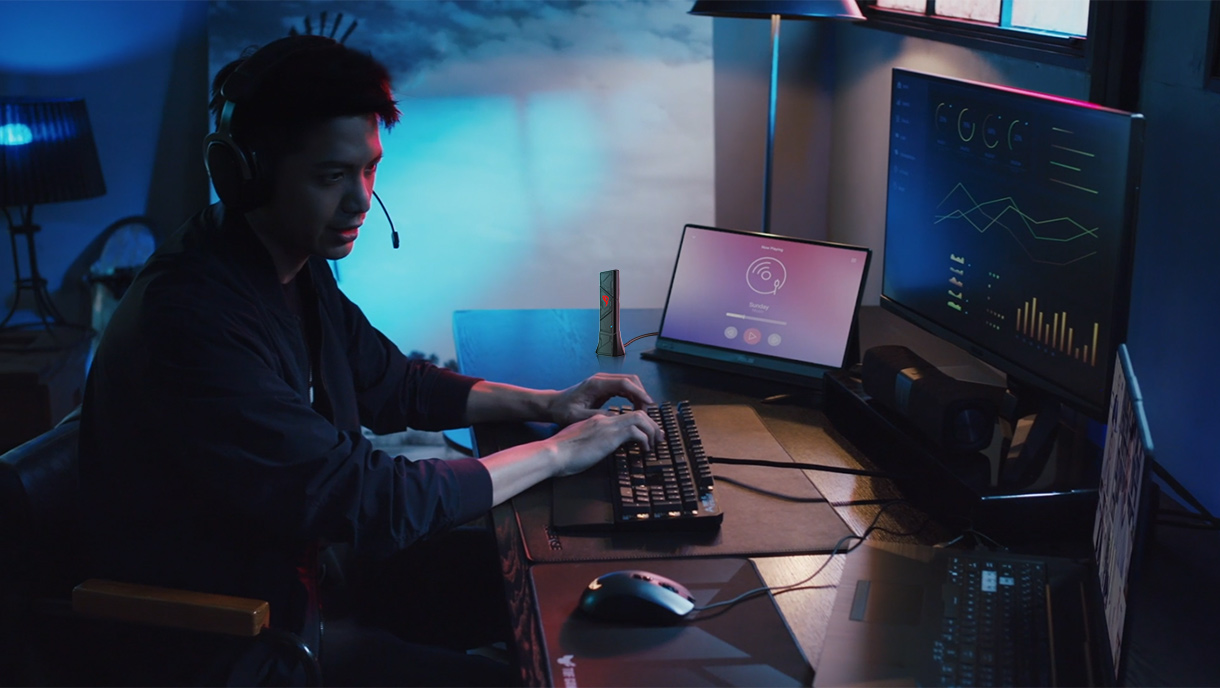 Un uomo con le cuffie è seduto a una scrivania con più monitor, che digita su una tastiera. La stanza è debolmente illuminata con sfumature blu.