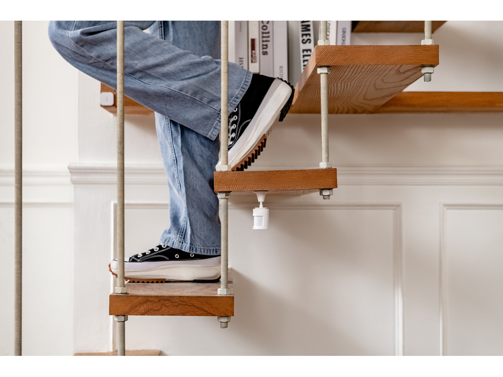 Beine und Füße einer Person auf Holztreppen. Sie trägt Jeans und schwarze Turnschuhe. Die Treppe hat Metallstützen und eine weiße Wand im Hintergrund.