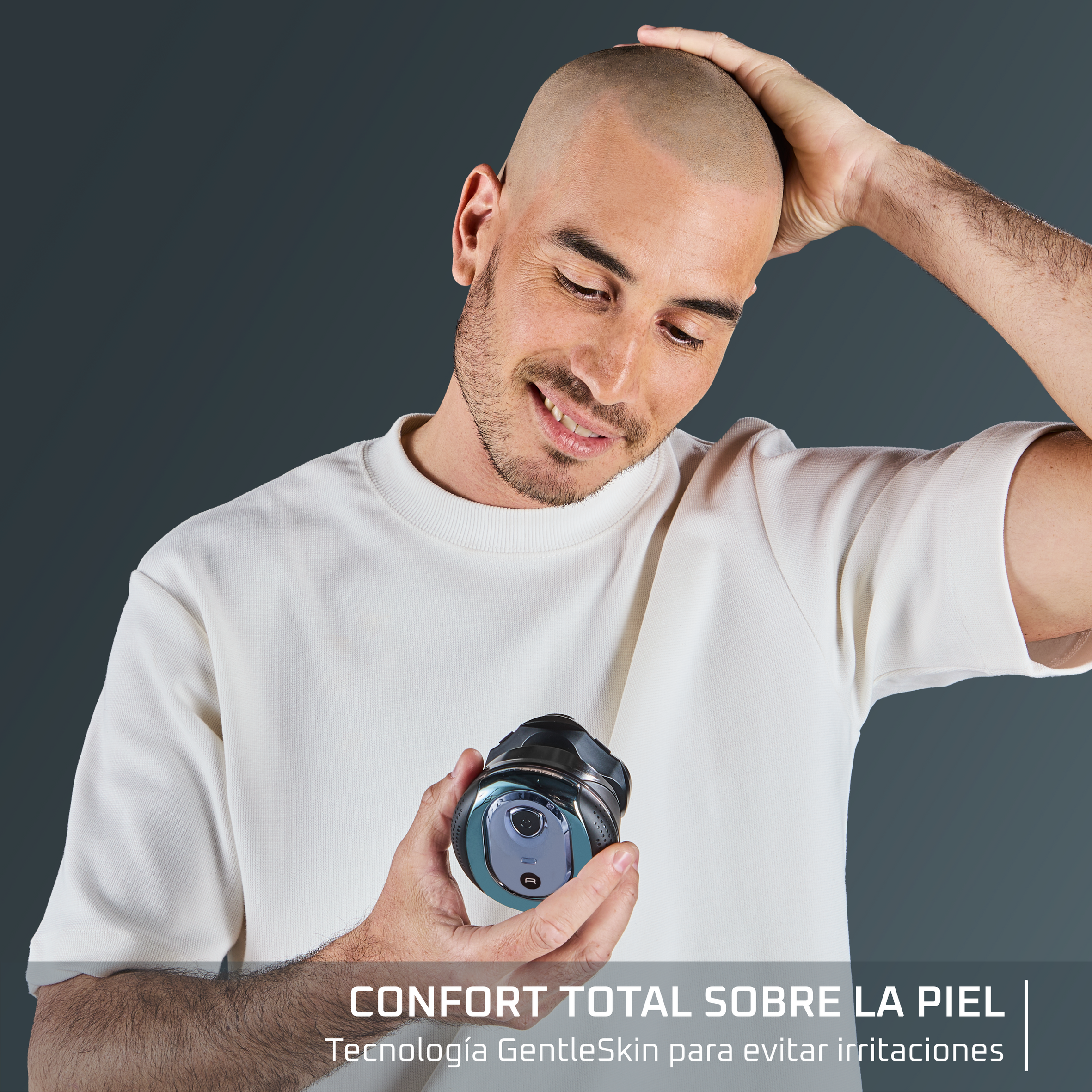 Un hombre sostiene una afeitadora, sonriendo. Se toca la cabeza. El fondo es gris.