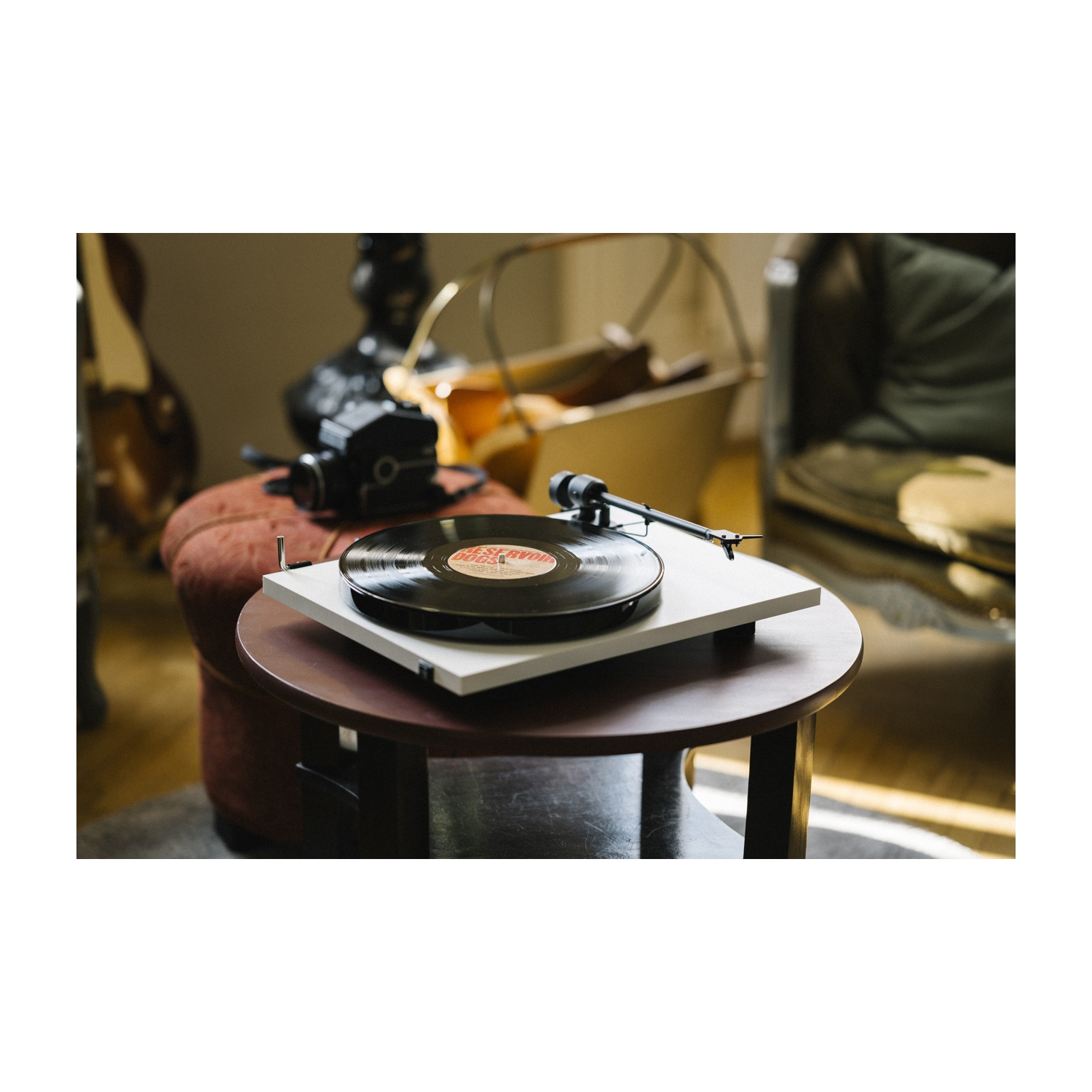 Un giradischi con un disco in vinile su un tavolino rotondo. Una fotocamera e uno sgabello accanto. Sfondo: un divano.