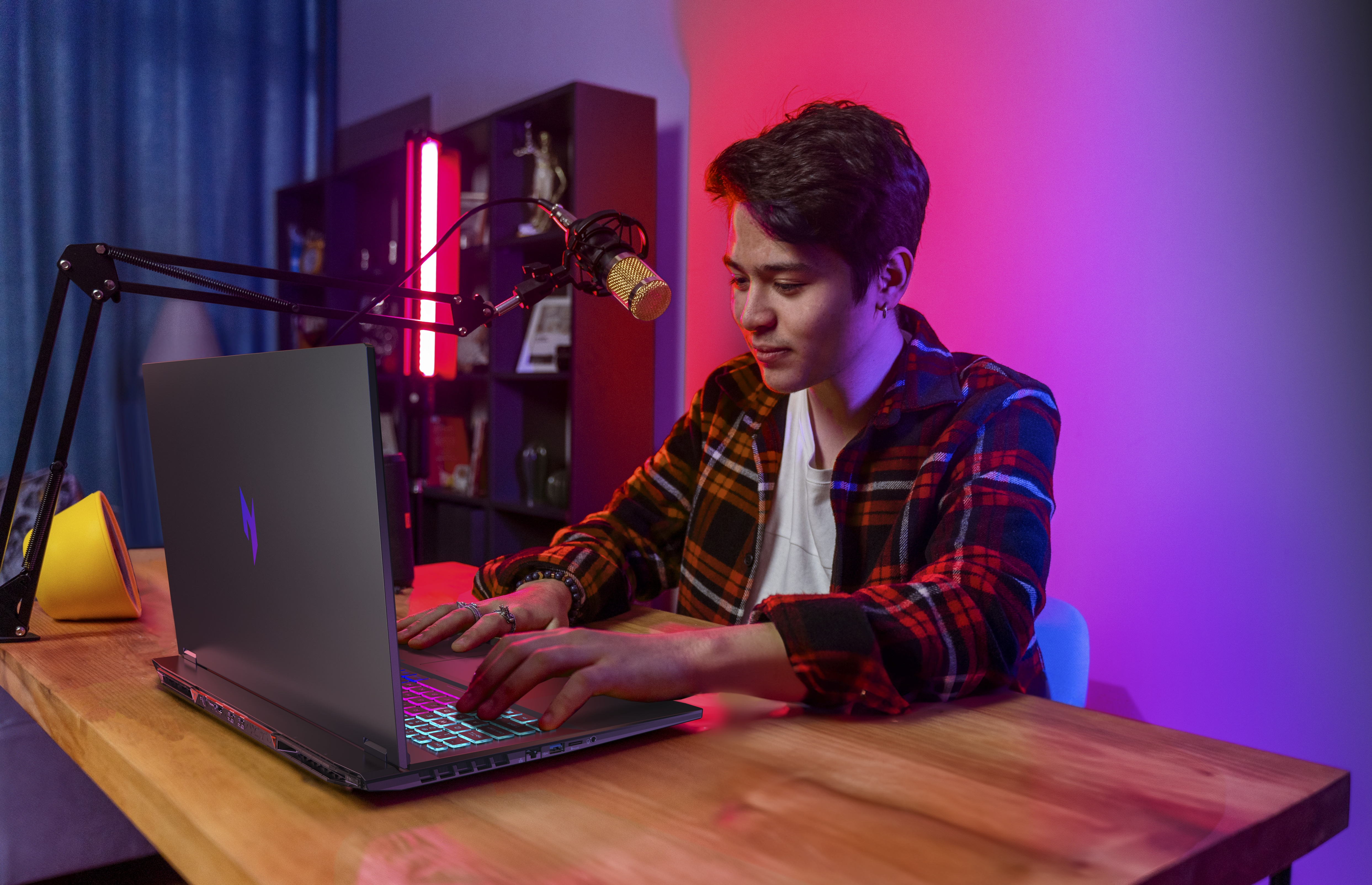 Ein junger Mann tippt an einem Schreibtisch auf einem Laptop. Ein Mikrofon ist vor ihm.