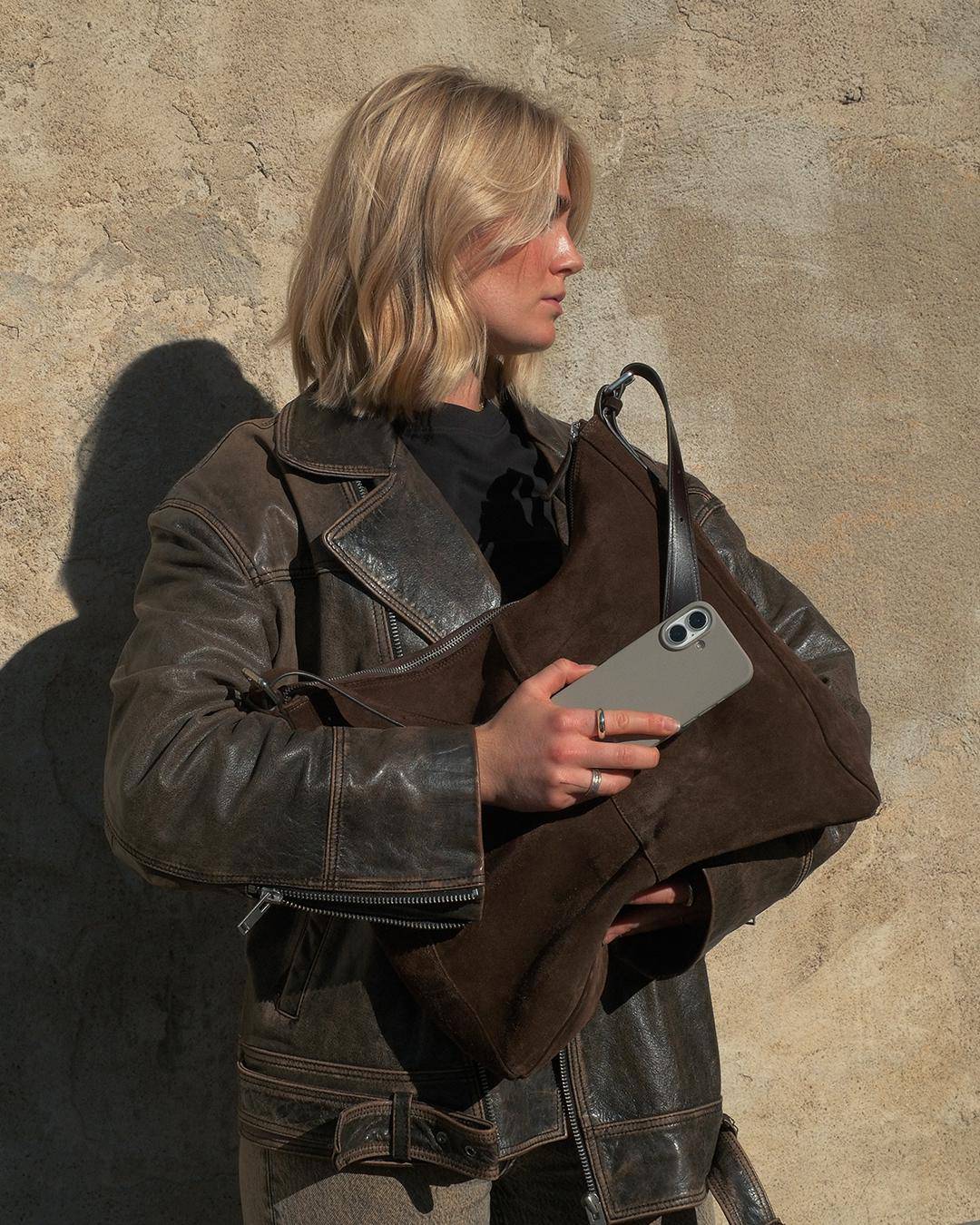 Blonde Frau in einer braunen Lederjacke hält eine braune Tasche und ein Telefon. Sie steht vor einer strukturierten Wand.