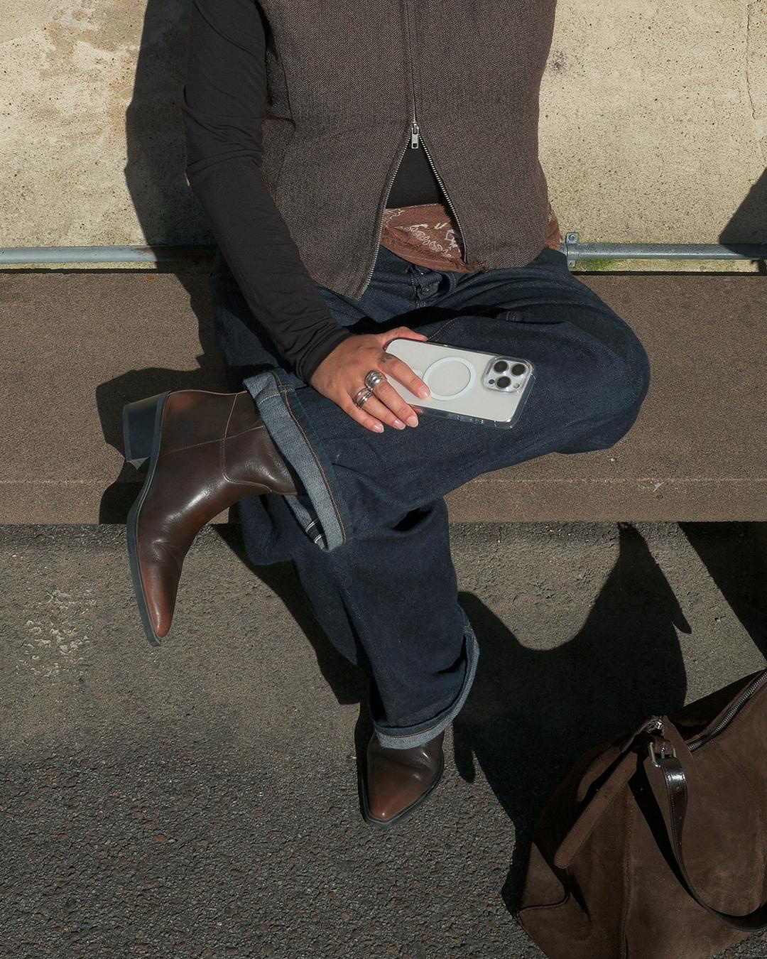 Person sitzt und hält ein Telefon. Trägt Jeans, Stiefel und eine Weste. Eine Tasche liegt daneben. Der Hintergrund ist eine Betonmauer.
