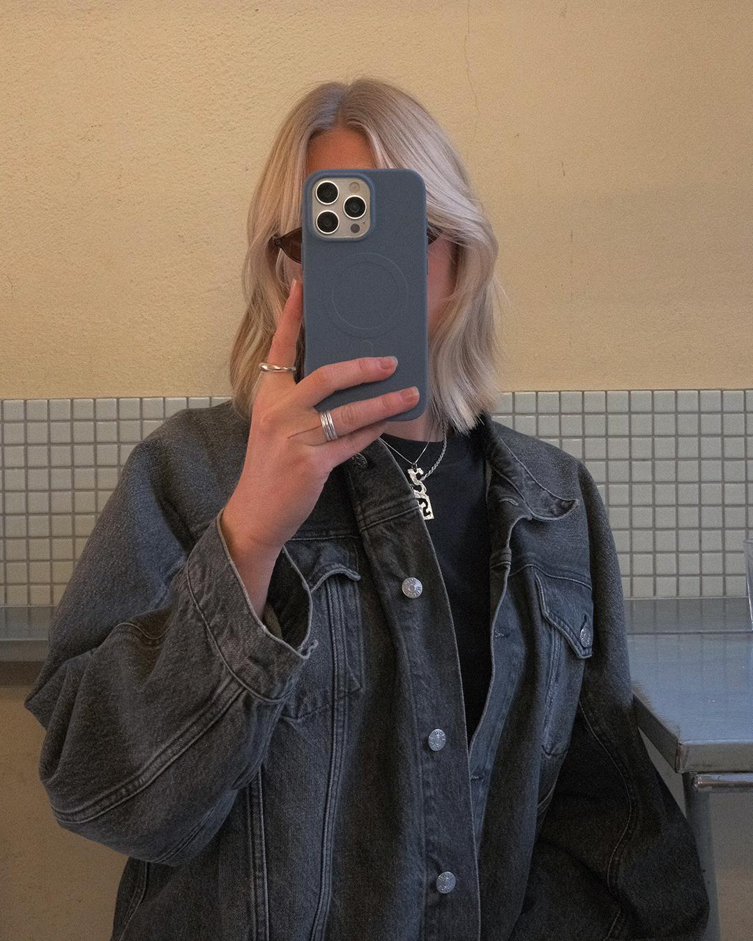 Person macht ein Selfie im Spiegel. Hält ein Telefon vor dem Gesicht. Trägt eine Jeansjacke. Blondes Haar, Schmuck und Fliesen im Hintergrund.