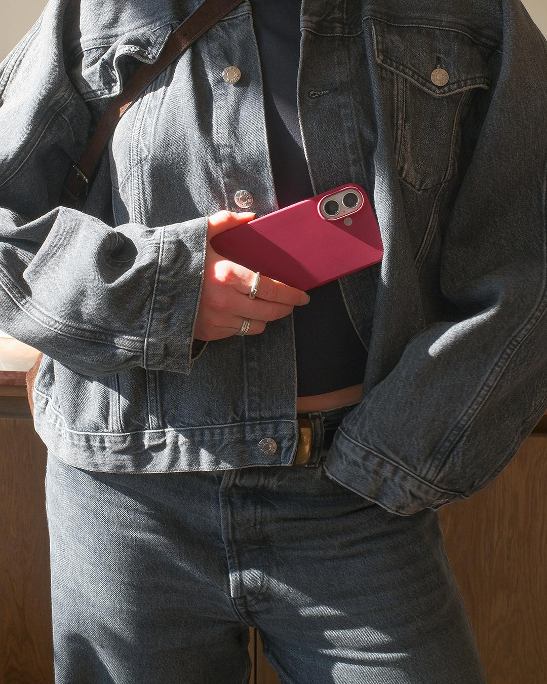 Person in Jeansjacke hält rotes Smartphone. Ringe und schwarzes Top. Jeans und braune Tasche sind sichtbar.