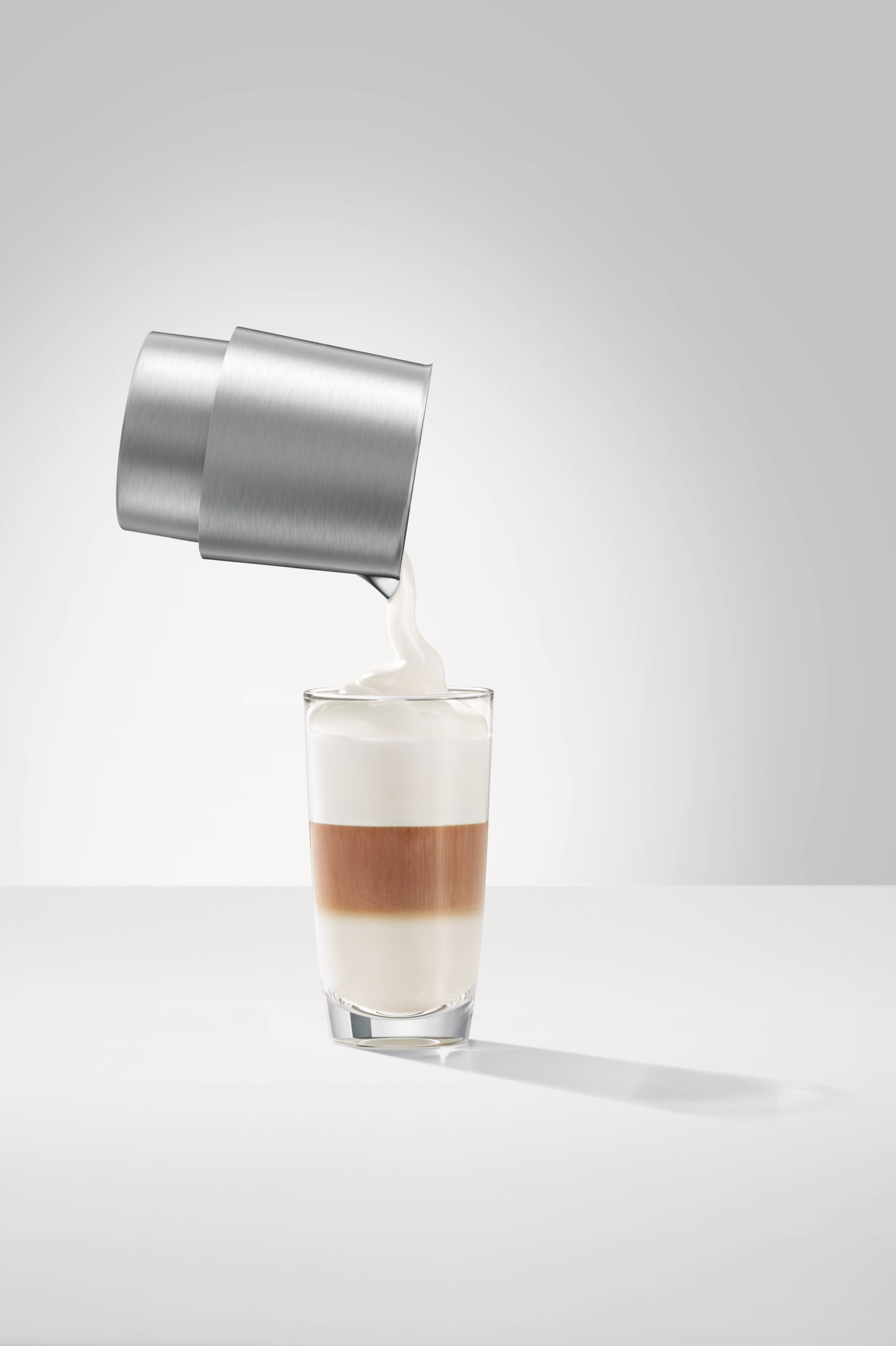 Ein Latte wird zubereitet. Ein Metallkrug gießt weißen Schaum in ein geschichtetes Kaffeeglas.