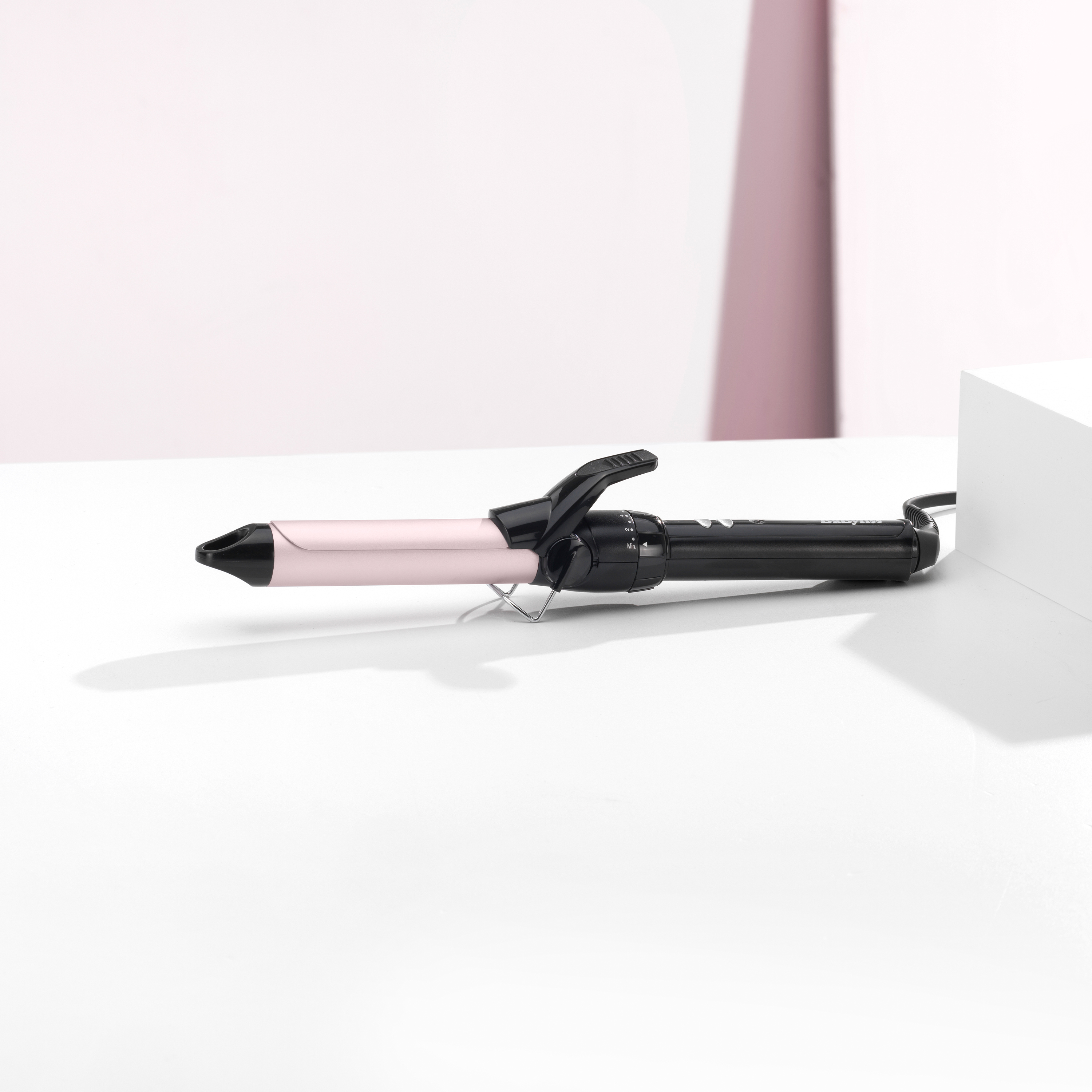 Ein Lockenstab auf einer weißen Oberfläche. Er hat einen rosa Schaft und einen schwarzen Griff. Der Hintergrund ist weiß und rosa.