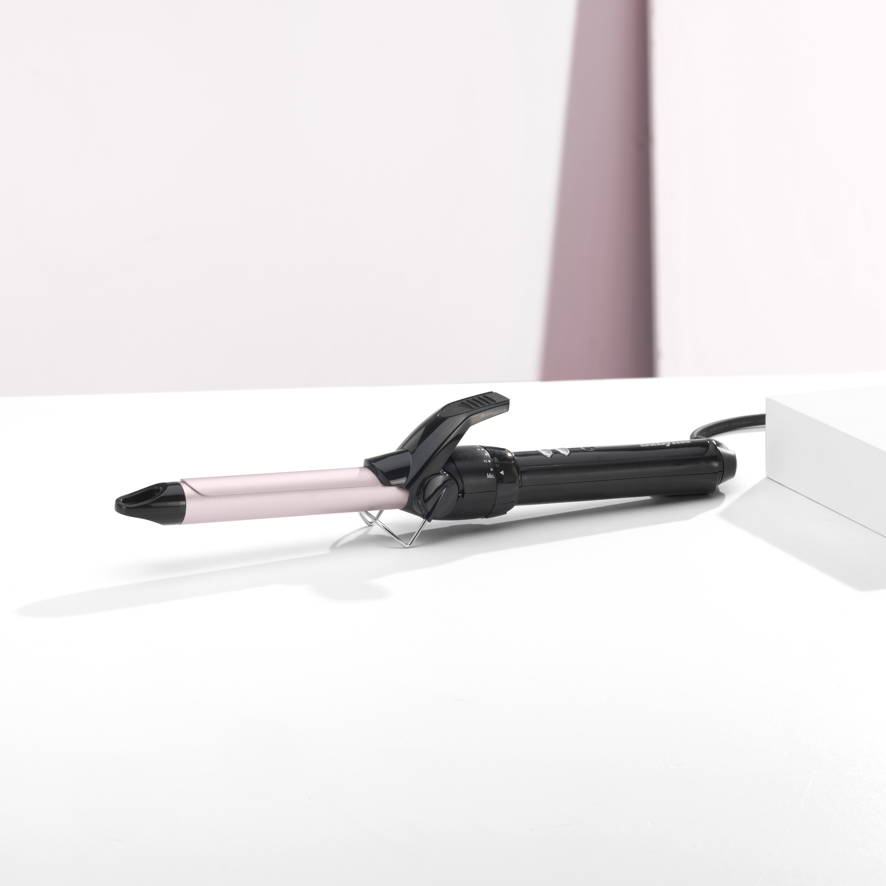 Ein Lockenstab liegt auf einer weißen Oberfläche. Er hat einen schwarzen Griff und einen rosa Zylinder. Der Hintergrund ist weiß.