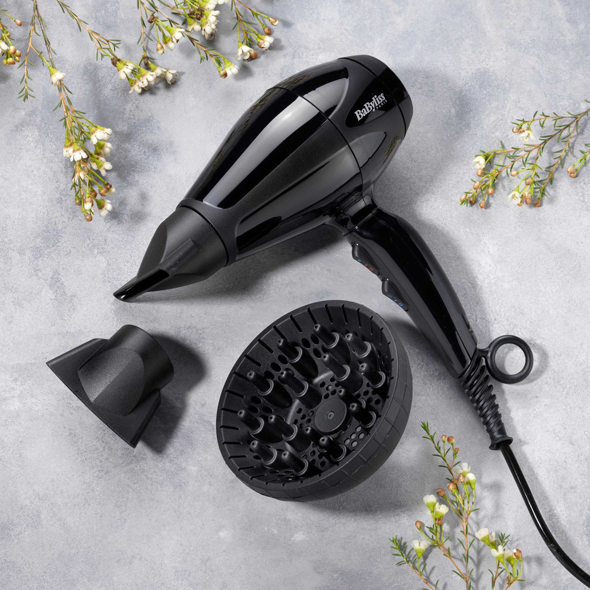 Ein schwarzer Haartrockner mit Aufsätzen und Blumen auf grauem Untergrund. Auf dem Trockner steht 'BaByliss'.