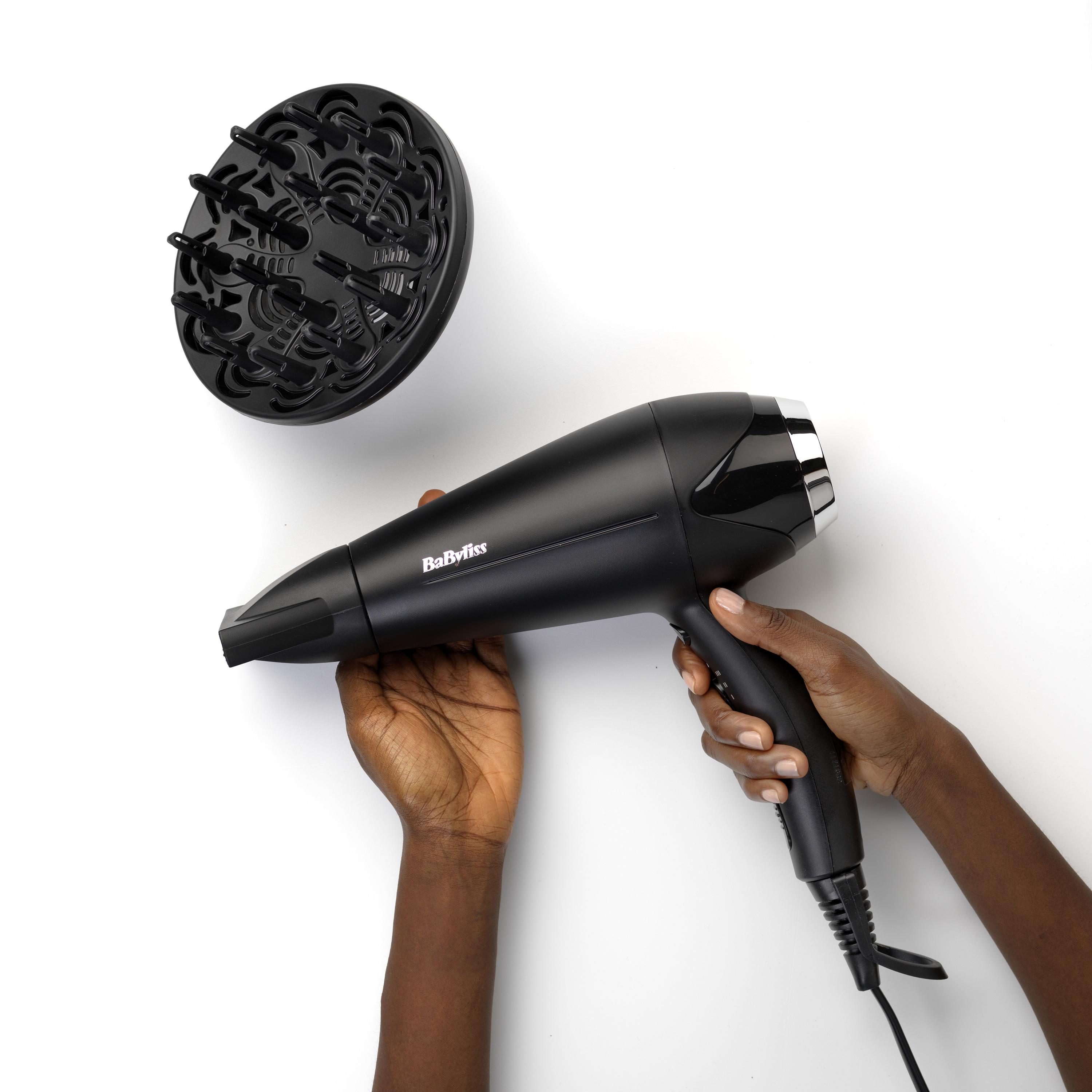 Schwarzer Haartrockner und Diffusor, gehalten von Händen auf weißem Hintergrund. Marke 'BaByliss'.