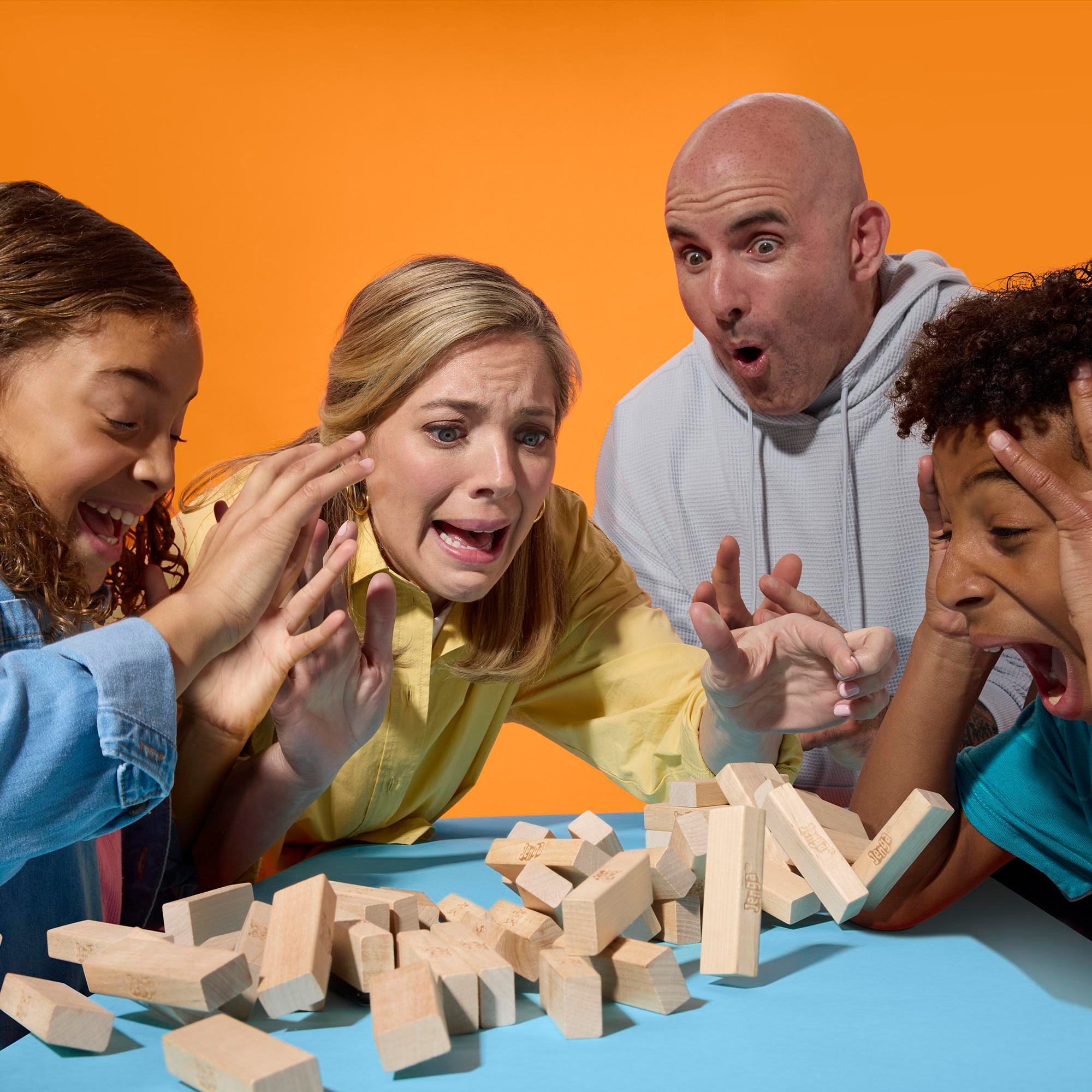 Familie spielt Jenga. Menschen sind schockiert, als der Turm einstürzt. Holzblöcke und blauer Tisch. Oranger Hintergrund.