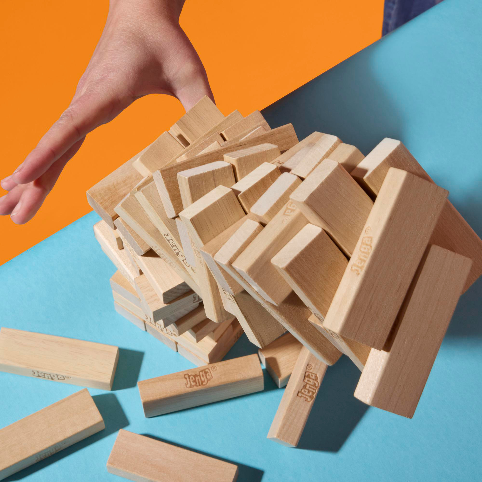 Eine Hand greift nach einem teilweise eingestürzten Jenga-Turm, mit verstreuten Blöcken auf blauem und orangefarbenem Hintergrund.