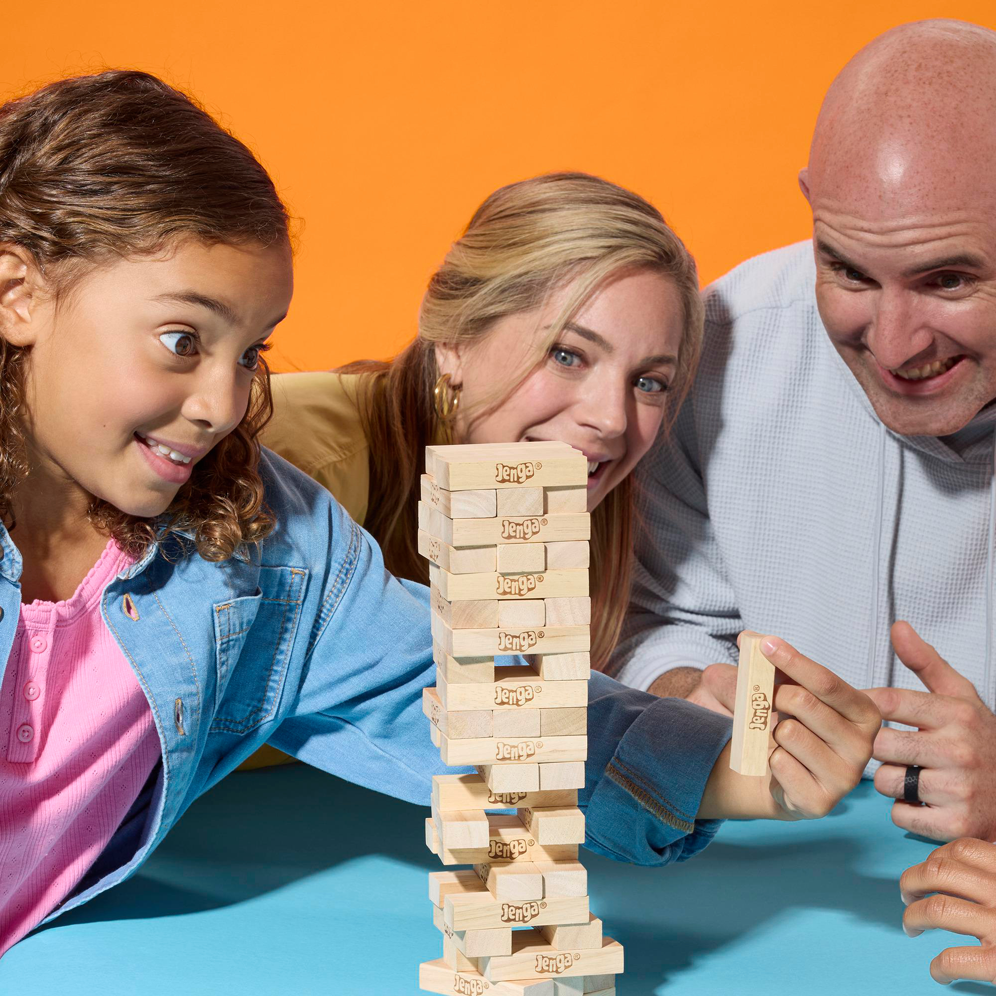 Drei Personen spielen Jenga. Ein Turm aus Blöcken steht auf einer blauen Oberfläche. Ängstliche Gesichter und ein Holzblock stehen im Fokus.