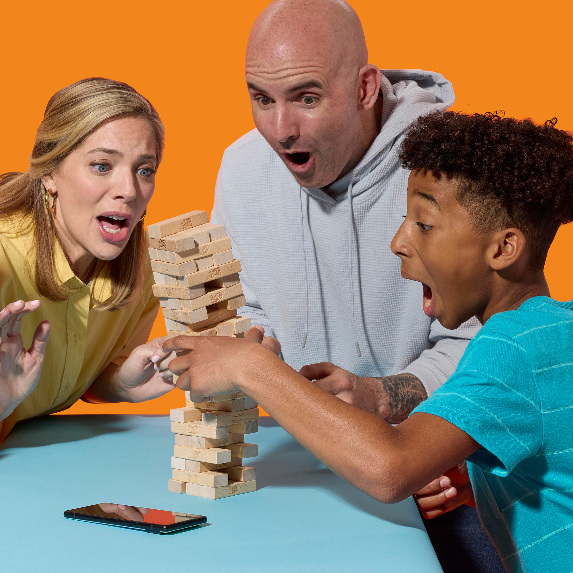 Drei Personen spielen Jenga, alle mit überraschten Ausdrücken. Ein Telefon liegt auf dem Tisch. Oranger Hintergrund.